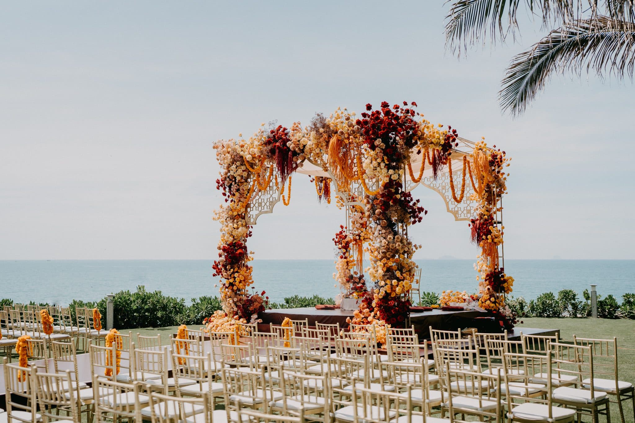 A floral mandap and rows of chairs set up on a lawn by the sea